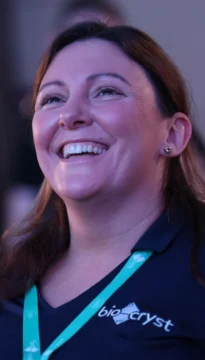 A woman with brown hair and a bright smile is wearing a blue shirt with "bio-cryst" logo and a teal lanyard.