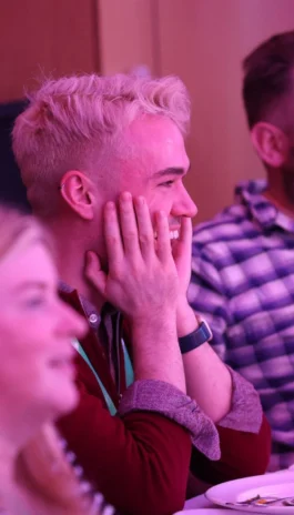 A person with short blond hair, wearing a dark shirt and smartwatch, is smiling with hands on their face while seated at a table during an event.