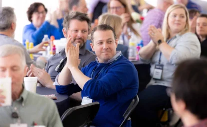 A group of people sit at tables, some applauding, during a gathering or event. They wear name tags and appear attentive.