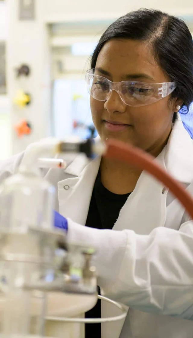 A person wearing safety goggles and a lab coat operates laboratory equipment in a scientific setting.