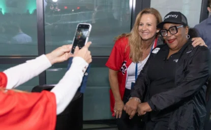 Two women pose for a photo while another person takes their picture with a smartphone, indoors. One woman is wearing a red shirt, and the other is dressed in black.