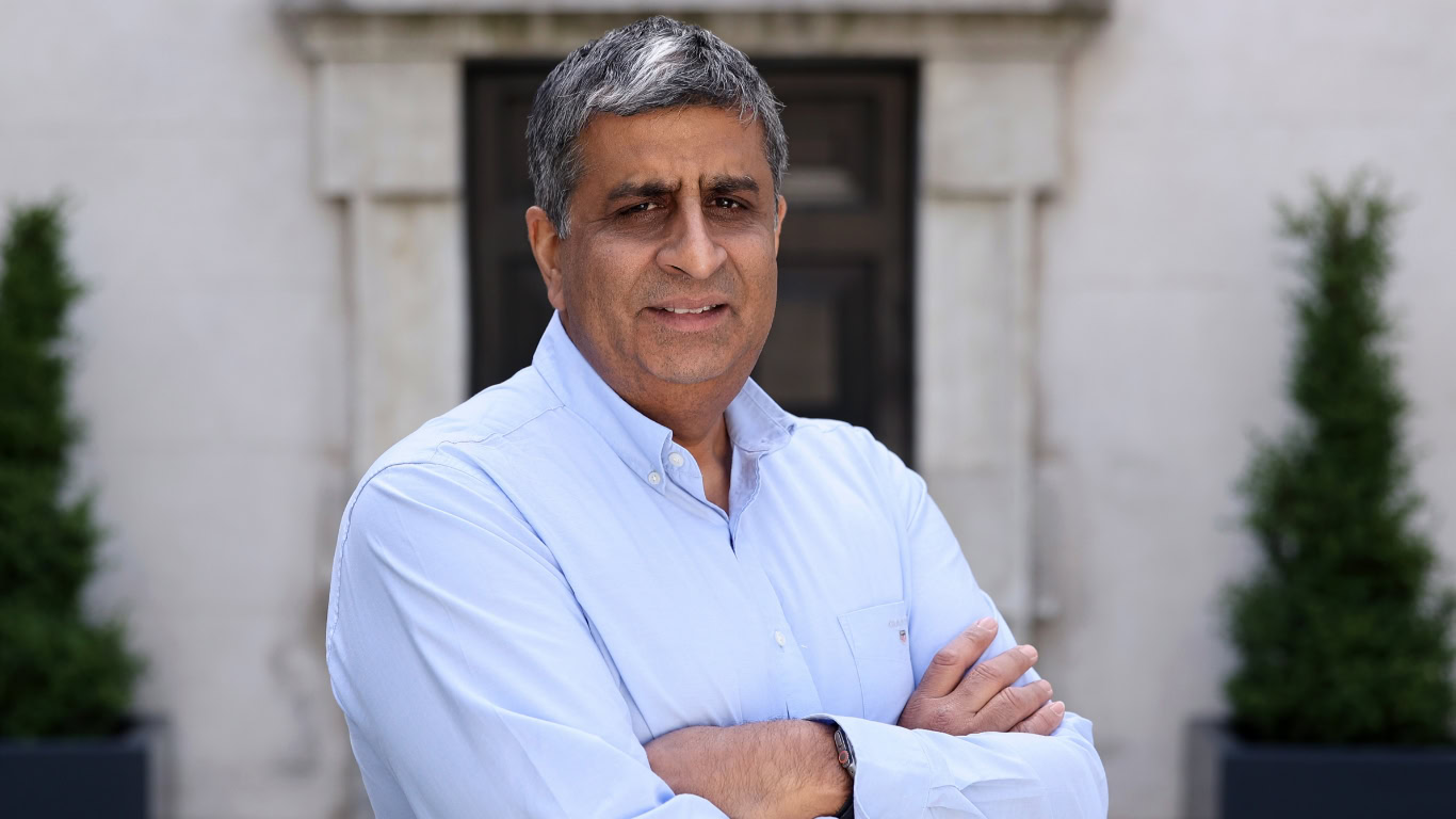 A man with gray hair stands outdoors with his arms crossed, wearing a light blue button-up shirt.